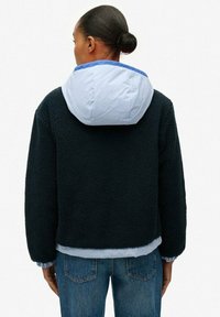 Persoon met donker haar in een bun, gekleed in een zwart fleecjacket met een lichtblauwe capuchon en blauwe jeans, draaiend met de rug naar de camera.