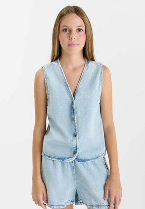 Jonge vrouw met steil lichtbruin haar, gekleed in een mouwloos lichtblauw denim vest met knopen en bijpassende denim shorts, staand tegen een witte achtergrond.