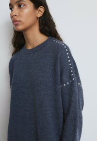 Joven con cabello oscuro y largo que lleva un suéter azul oscuro con tachuelas plateadas a lo largo de las costuras del hombro y la manga, mirando hacia la izquierda.