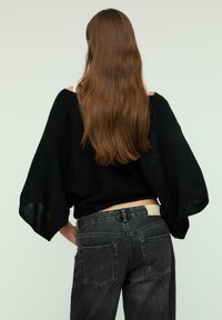 Pull en tricot noir avec des manches larges, associé à un jean taille haute gris foncé avec poches arrière et une étiquette sur la taille.