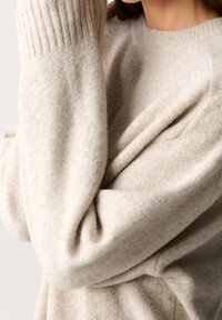 Pull en laine beige avec des poignets côtelés et un col rond, présentant un matériau doux et texturé ainsi qu'un tissu drapé pour un ajustement décontracté.