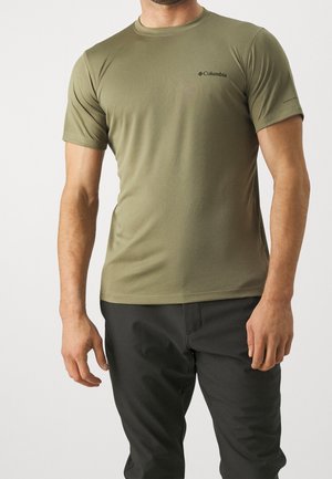 Mężczyzna ubrany w oliwkowo-zielony t-shirt Columbia z krótkim rękawem i czarne spodnie, stojący na jednolitym jasnym tle.