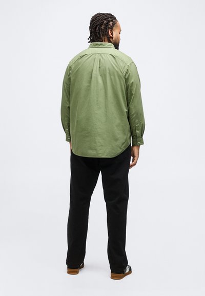 Άνδρας με πλεξούδες, που φοράει πράσινο μακρυμάνικο πουκάμισο, μαύρο παντελόνι και μαύρα και λευκά παπούτσια, στέκεται γυρισμένος με την πλάτη σε απλό φόντο.