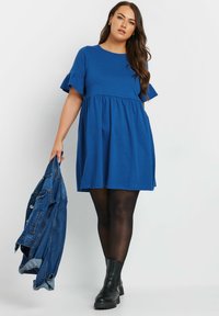 Kort blå klänning med korta volangärmar, en rynkad midja och en utställd kjol; matchad med svarta ankelskoboots och håller en jeansjacka.