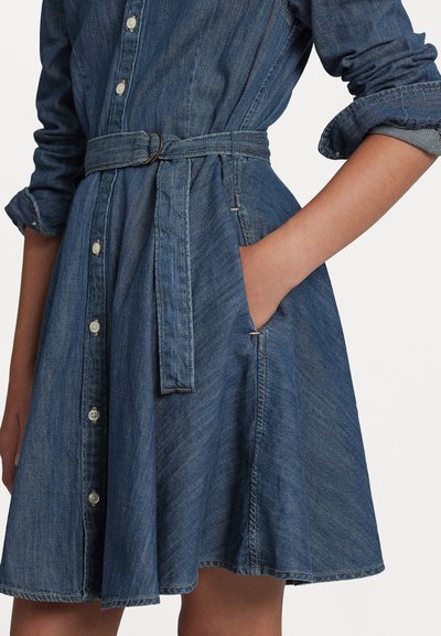 Denim skjortekjole i mørkeblå, med en innsnevret midje med belte, lange ermer med oppbrettede mansjetter og sidelommer.