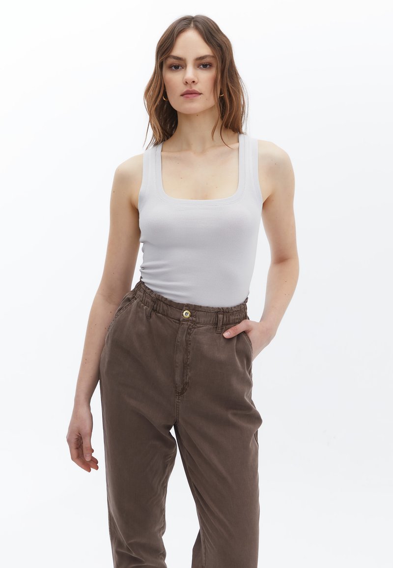 Graues geripptes Tanktop mit quadratischem Ausschnitt, kombiniert mit braunen Hose mit elastischem Bund und einer Knopfdetails vorne. Einheitlicher Hintergrund.