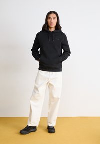 Felpa nera con cappuccio, polsini e tasca anteriore, abbinata a pantaloni bianchi a gamba larga e sneakers nere testurizzate. Outfit semplice e casual.