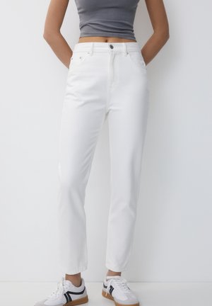 Jean blanc slim-fit porté avec un débardeur court gris et des baskets blanches avec des détails noirs et marron.