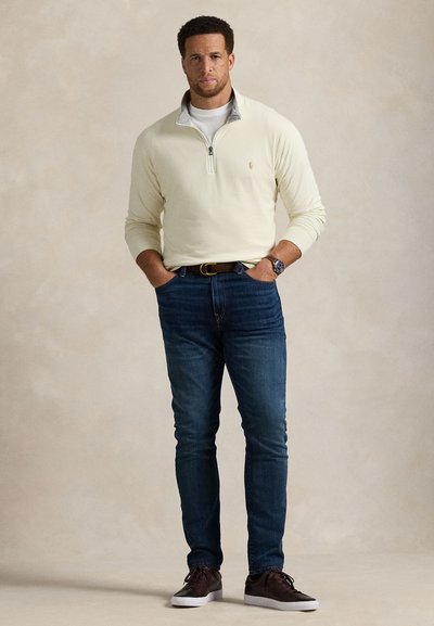 Ανοιχτό κίτρινο πουλόβερ με φερμουάρ μέχρι τη μέση και γκρι λεπτομέρειες, σκούρο μπλε τζιν παντελόνι και καφέ αθλητικά παπούτσια. Διαθέτει μικρό λογότυπο στο στήθος.