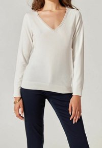 Kremowy sweter z długim rękawem i dekoltem w serek, wykonany z gładkiej tkaniny, z prążkowanym wykończeniem na dole i mankietach, zestawiony z granatowymi spodniami.