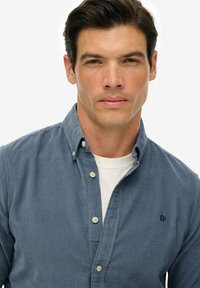 Uomo con capelli scuri che indossa una camicia blu con bottoni sopra una maglietta bianca, che guarda dritto di fronte a sé con un'espressione neutra.