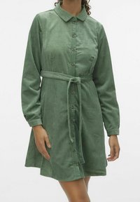 Žena v zelených kordových šatech na knoflíky s dlouhými rukávy a uvázaným páskem v pase, stojící na pozadí jednoduché bílé stěny.