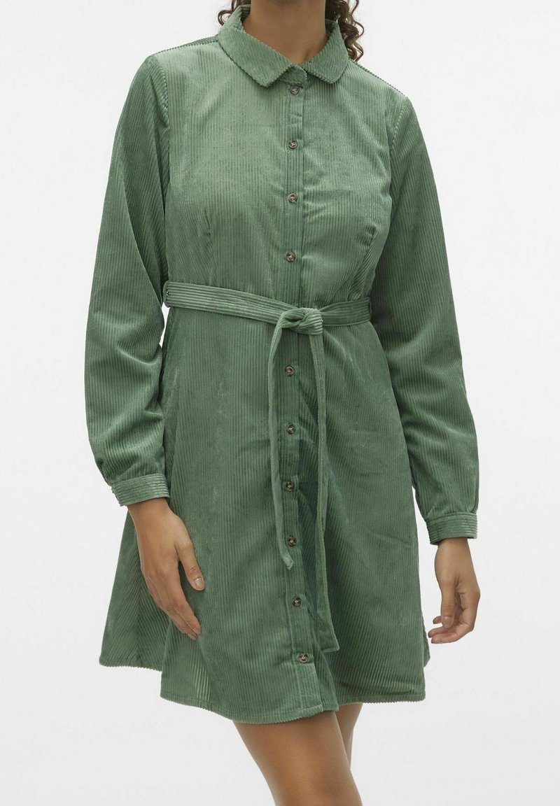 Žena v zelených kordových šatech na knoflíky s dlouhými rukávy a uvázaným páskem v pase, stojící na pozadí jednoduché bílé stěny.