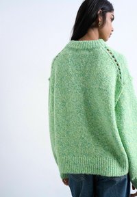 Femme aux longs cheveux noirs portant un pull ample en maille vert clair avec des détails en trous sur les manches et un jean bleu en denim, vue de profil.