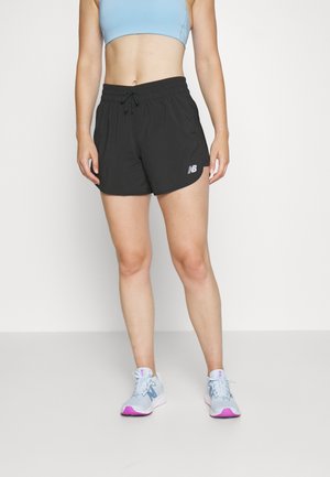 Vrouw draagt zwarte New Balance shorts, lichtblauwe sportbeha en lichtblauwe en roze hardloopschoenen, staand tegen een effen witte achtergrond.