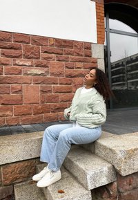 Young woman with curly hair wearing a chunky knit sweater and jeans sits on outdoor stone steps against a red brick wall.