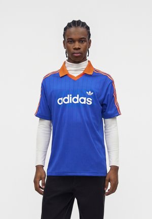 Nuori mies punotut hiukset, joka pukeutunut siniseen Adidas-paitaan, jossa on oranssi kaulus ja raidat, kerroksittain valkoisen pitkähihaisen paidan päällä.