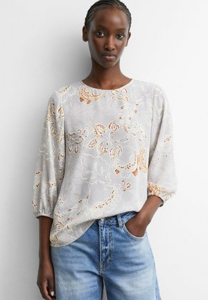 Junge Frau mit geflochtenem Haar, die eine hellfarbene Bluse mit Blumenmuster und blaue Jeans trägt, mit einer Hand in der Tasche, steht vor einer weißen Wand.