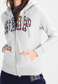 Sweat à capuche gris zippé avec une poche kangourou à l'avant. Présente un grand logo "GAP" en rouge, blanc et bleu avec un design texturé. Texture de tissu douce.