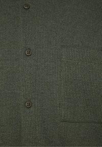 Gros plan sur un tissu tricoté côtelé vert foncé avec trois boutons marron et une petite poche sur le côté droit.