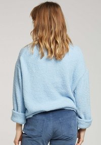 Pull oversized bleu clair avec une finition texturée et duveteuse. Il présente des poignets roulés et une coupe décontractée, associé à un pantalon en velours côtelé bleu foncé.