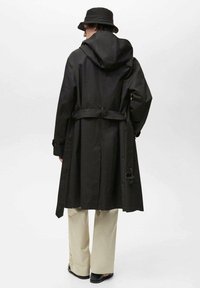 Trench coat negru cu glugă, talie strânsă și mâneci lungi. Asortat cu pantaloni bej cu picioare largi și o pălărie neagră tip bucket.