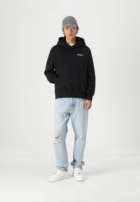 Felpa nera con cappuccio e logo bianco "LEVI'S", abbinata a jeans strappati blu chiaro e sneakers bianche. Un cappellino da baseball grigio completa il look.