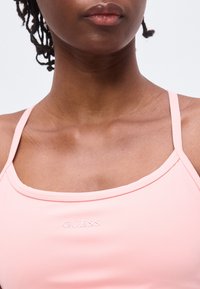 Κοντινό πλάνο ενός ατόμου που φοράει ανοιχτόροζ τοπ με λεπτές τιράντες και το λογότυπο "GUESS" στο στήθος.