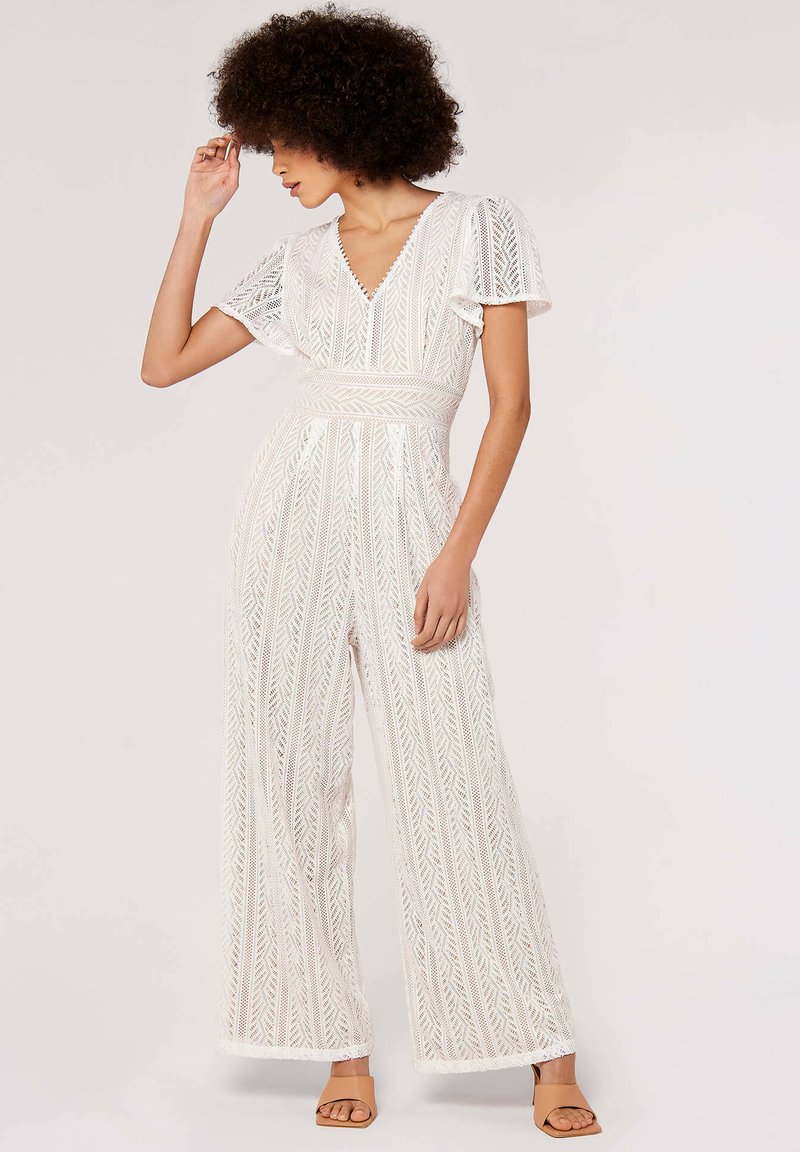 White lace jumpsuit with vertical patterns, V-neckline, short flutter sleeves, and wide leg design. Worn with tan block-heeled sandals.