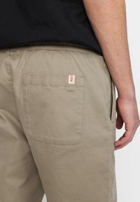 Beige cargo shorts met een zijzak en een achterzak met een klein rood label. Gemaakt van een lichtgewicht, duurzame stof.