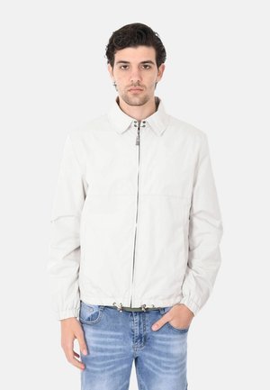 Giovane uomo con capelli scuri che indossa una giacca bianca con cerniera e jeans blu, in piedi davanti a uno sfondo bianco semplice.