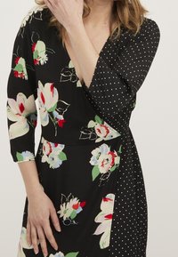 Černé šaty na zavazování s květinovými vzory v červené, růžové a zelené barvě, s puntíkovaným vzorem na jednom rukávu. Vyrobeno z měkké tkaniny s hladkým povrchem.