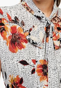 Blusa floreale con un laccio al collo, realizzata in tessuto leggero. Predominantemente bianca con strisce nere a texture e vivaci fiori arancioni.