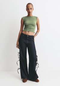 Zelený žebrovaný crop top a tmavé denimové široké kalhoty s postranními šňůrkami. Modelka má na sobě hnědou obuv a stříbrný náramek. Minimalistické pozadí.