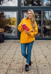 Red leather wallet with a clasp, held by a person in a bright yellow oversized sweater and blue jeans, standing on a textured stone surface.