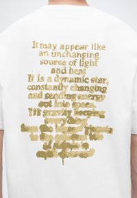 Chemise blanche avec un texte doré dans le dos décrivant le soleil comme une étoile dynamique et changeante émettant de l'énergie dans l'espace.