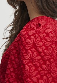 Haut rouge brodé avec des motifs floraux, en tissu texturé et à col rond, mettant en avant des détails raffinés et une silhouette structurée.