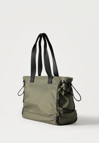 Τσάντα ώμου σε χρώμα ελιάς με μαύρες λαβές και ρυθμιζόμενα μαύρα κορδόνια στα πλαϊνά, τοποθετημένη σε απλό ανοιχτόχρωμο φόντο.