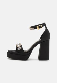Sandali platform neri con tacco spesso, finitura satinata e strass decorativi sulla fascia anteriore. Fibbia dorata sulla cinghia alla caviglia.