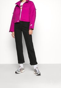 Fuksjowa pikowana kurtka z wysokim kołnierzem, czarne proste jeansy oraz srebrne sneakersy z białymi akcentami. Widoczne białe skarpetki.