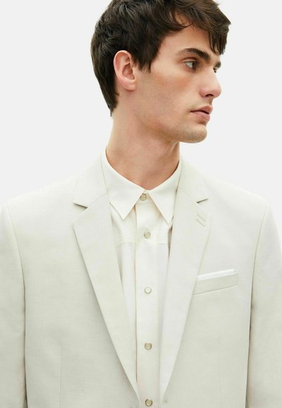 Giacca da abito beige chiaro con design a un petto, rever a scollo e taschino sul petto. Sotto, una camicia crema con bottoni visibili.