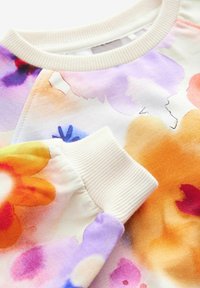 Gros plan sur un vêtement avec un col et des poignets côtelés couleur crème, présentant un motif floral aquarelle coloré en nuances d'orange, violet, jaune et rouge.