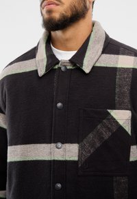 Černá a šedá kostkovaná bunda se světle zelenými akcenty, zapínání na knoflíky, špičatý límec a náprsní kapsa s diagonálními vzory.
