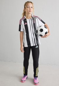 Juventus zīmola melnbalta svītrainā krekls ar rozā akcentiem, melnas sporta bikses ar dzeltenām svītrām un rozā futbola apaviem. Tur rokās futbola bumbu.