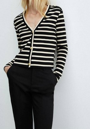 Schwarz-weiße gestreifte Strickjacke mit V-Ausschnitt, ausgestattet mit goldenen Knöpfen und langen Ärmeln, kombiniert mit schwarzen Hosen. Glatte Stofftextur.