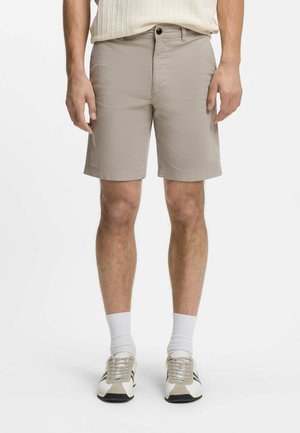 Mann trägt beige maßgeschneiderte Shorts, weiße gerippte Socken und weiße Turnschuhe mit schwarzen Streifen, steht vor weißem Hintergrund.