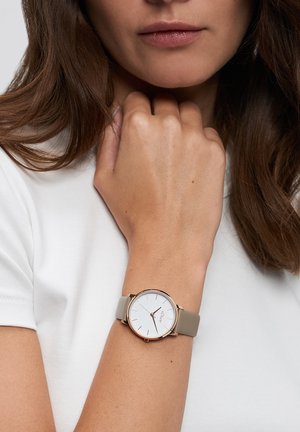 Runde Uhr mit weißem Zifferblatt, goldfarbenen Akzenten und einem hellgrauen Silikonarmband, getragen an einem Handgelenk, das ein schlichtes weißes Hemd trägt.