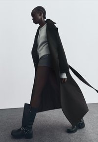 Kvinna går klädd i en lång svart kappa över en grå tröja, svarta shorts och svarta stövlar, på en grå mattlagd golv mot en vit bakgrund.