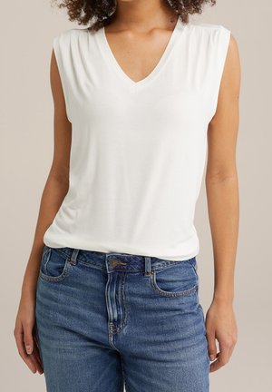 Kobieta w białym, bezrękawnikowym topie z dekoltem w serek, wpuszczonym w niebieskie dżinsy, na jednolitym tle.