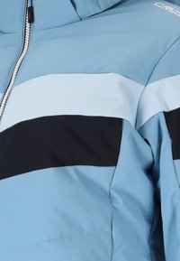 Lys blå jakke med horisontale striber i mørkeblå og hvid. Har en frontlynlås og en blød, struktureret overflade.